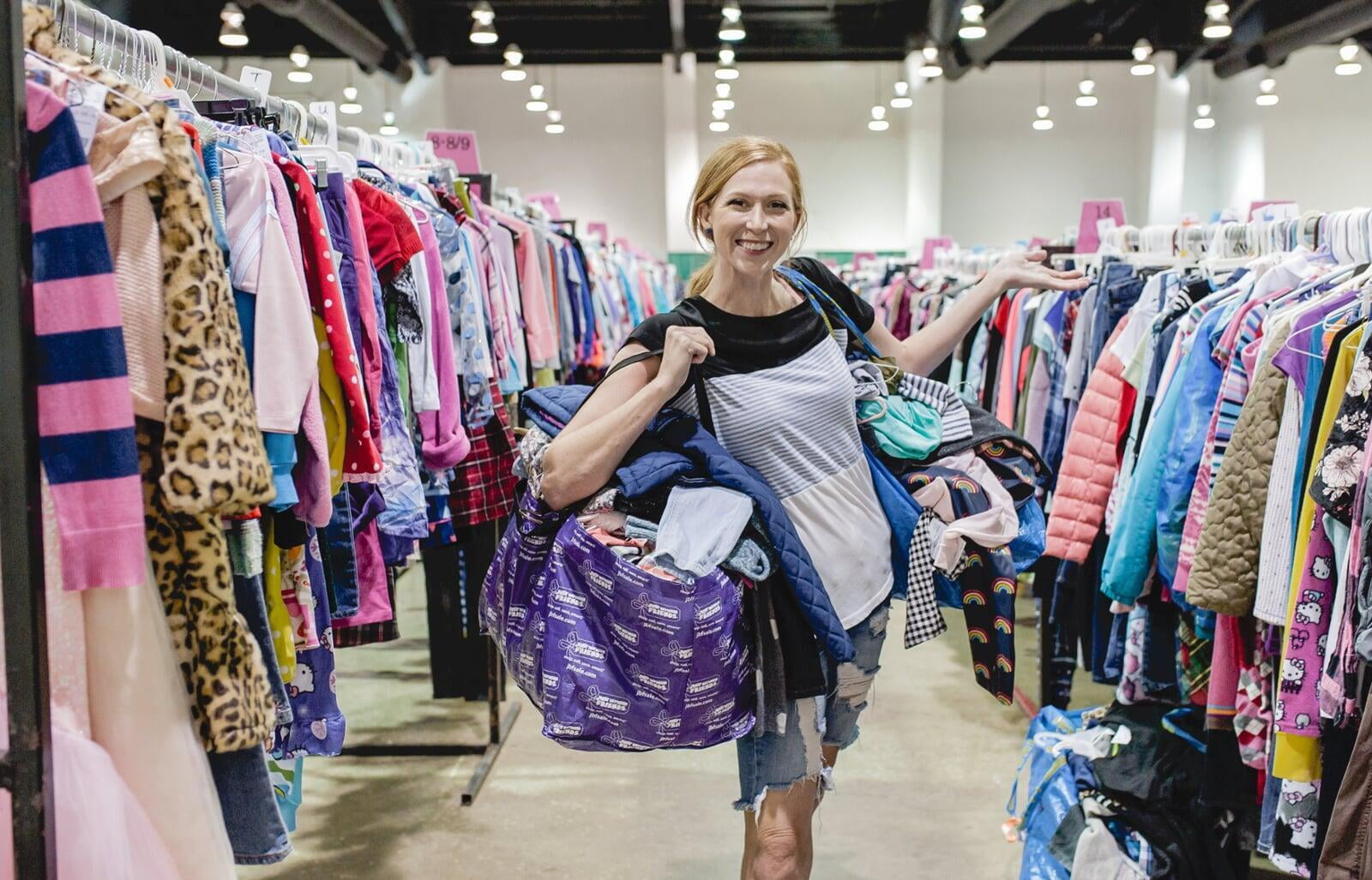 Mom holds hand up to show of rows and rows of clothing for sale at 50 to 90% off retail prices! She is carrying two bags of items that she is going to purchase.