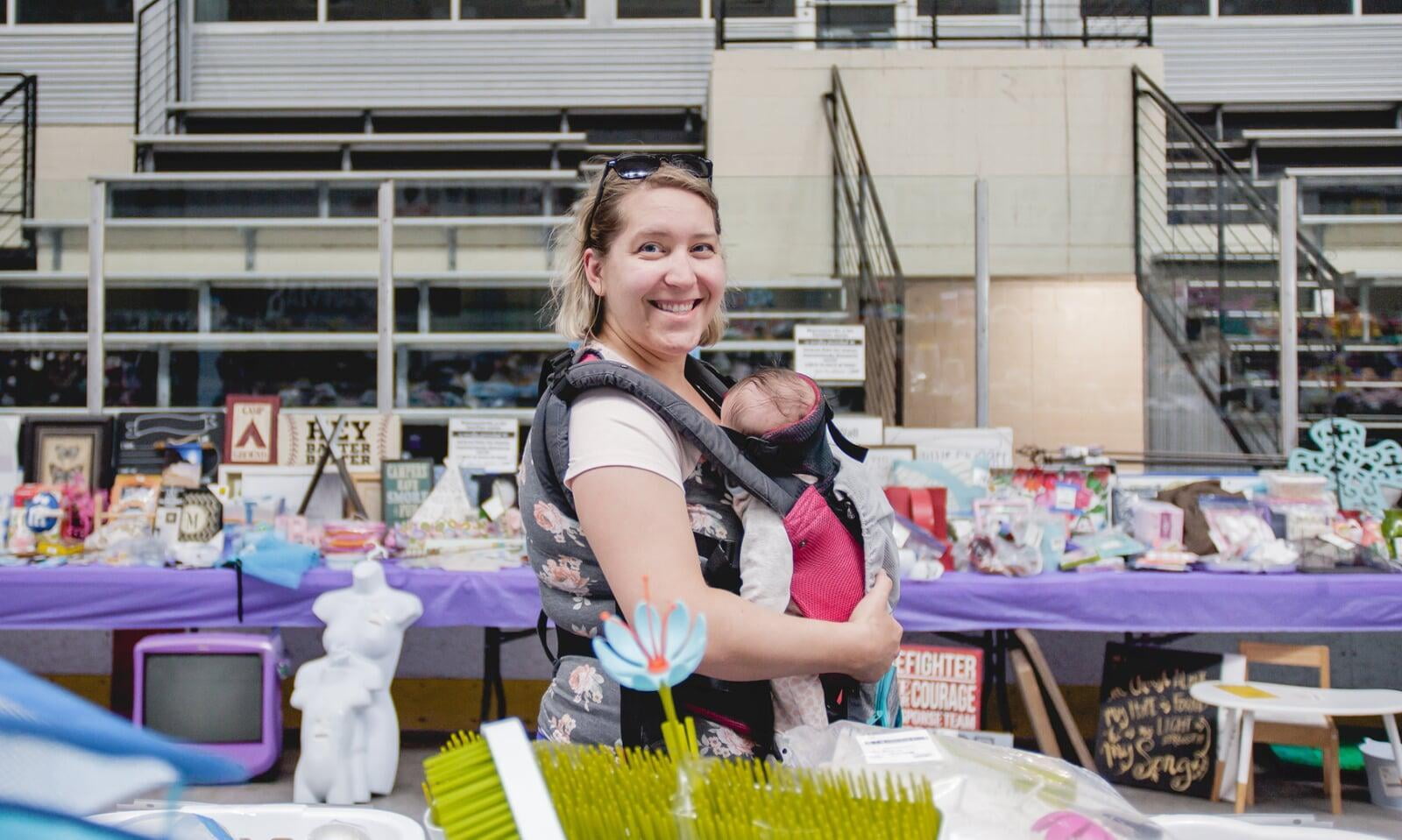 Mom smiles as she carries her baby in a front-pack carrier. She is walking past tables covered with items for sale at Just Between Friends.