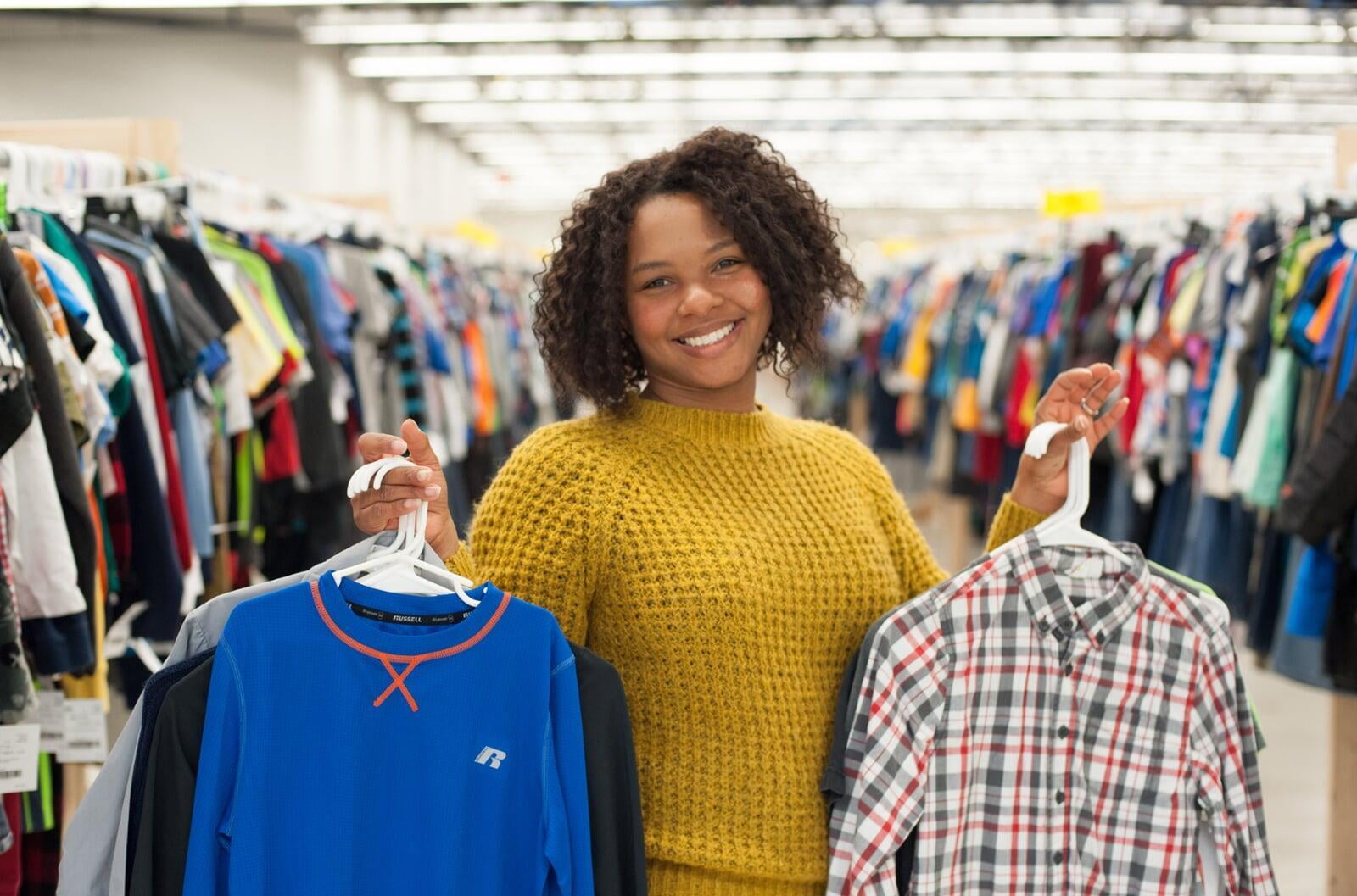 A smiling woman hold up boys long sleeved shirts on hangers. Behind her there are racks of boys clothing.